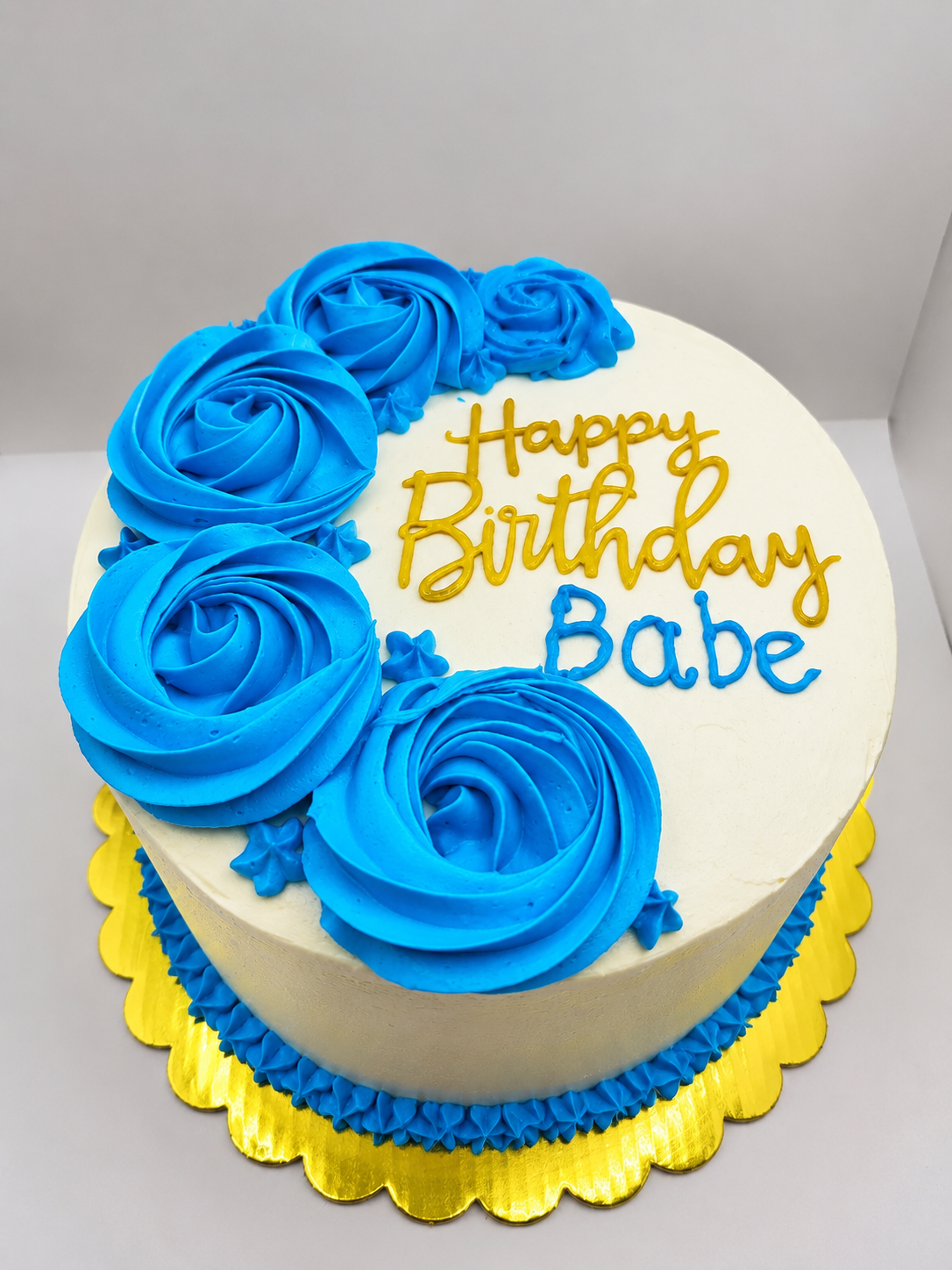 White whipped cream cake with a cascade of bright blue buttercream rosettes, piped star border and gold and blue lettering